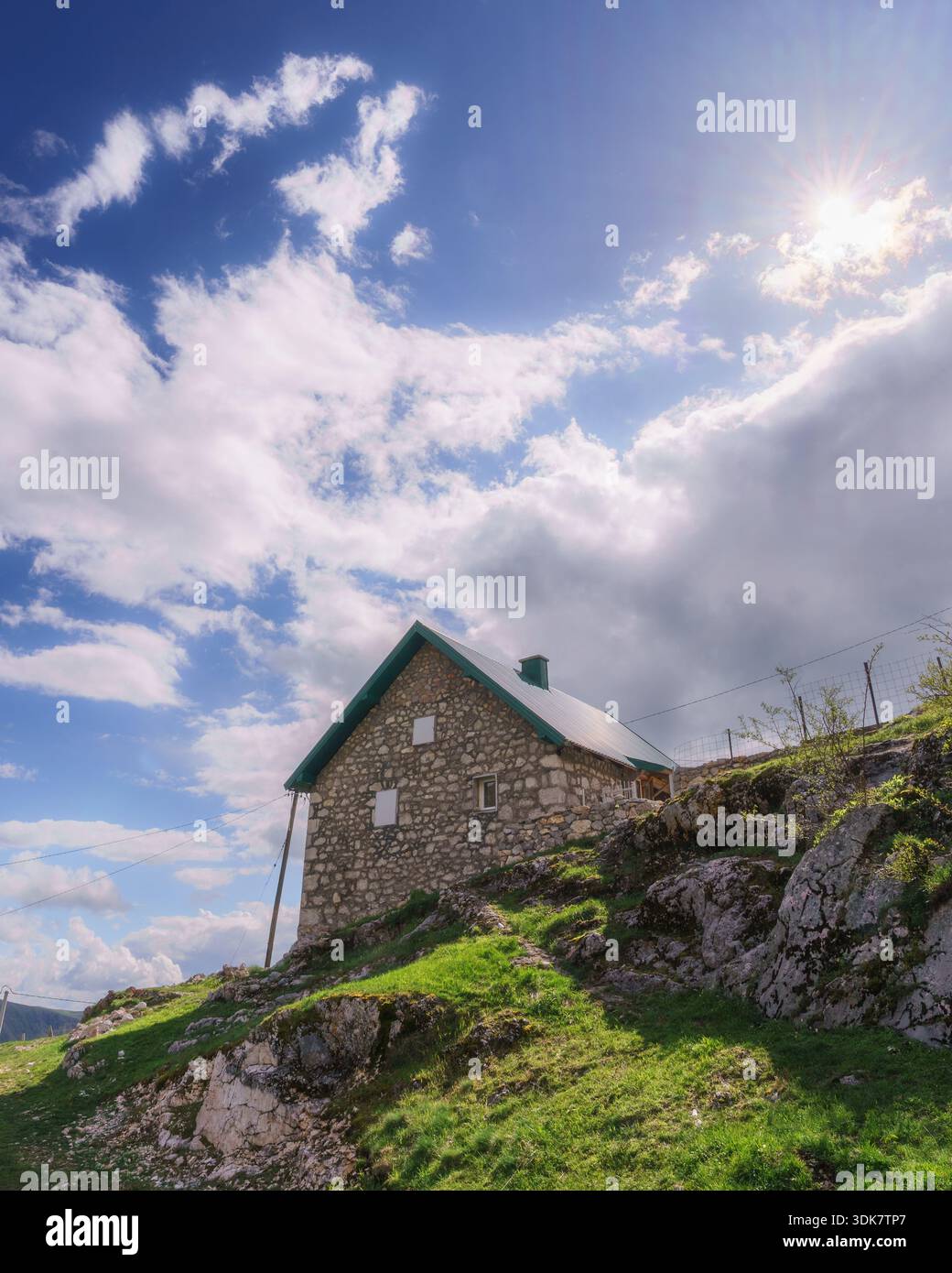 Blick auf eine Steinhütte auf einem grasbewachsenen Hügel unter hellem Himmel mit verstreuten Wolken und strahlender Sonne, Lukomir, Föderation von Bosnien und Herzegowina Stockfoto