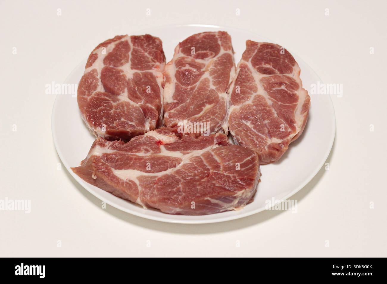 Blick von oben auf rohe Schweinefleisch-Steaks auf einem weißen Teller, zubereitet zum Kochen auf einem sauberen Küchentisch Stockfoto