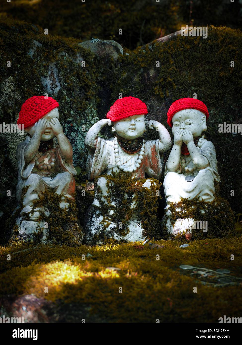 Drei Steinstatuen in einem Wald im Motiv „See no Evil, hör no Evil, sprich no Evil“ mit roten Kappen. Stockfoto