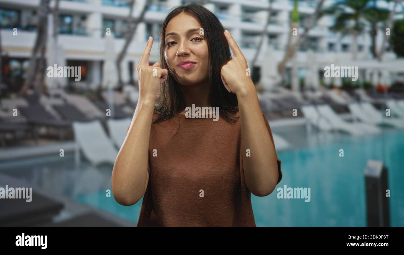 Die hispanische Frau zeigt mit beiden Zeigefingern nach oben auf die Liegestühle am Außenpool des Hotels, während sie lächelt; verspieltes Selbstvertrauen. Stockfoto