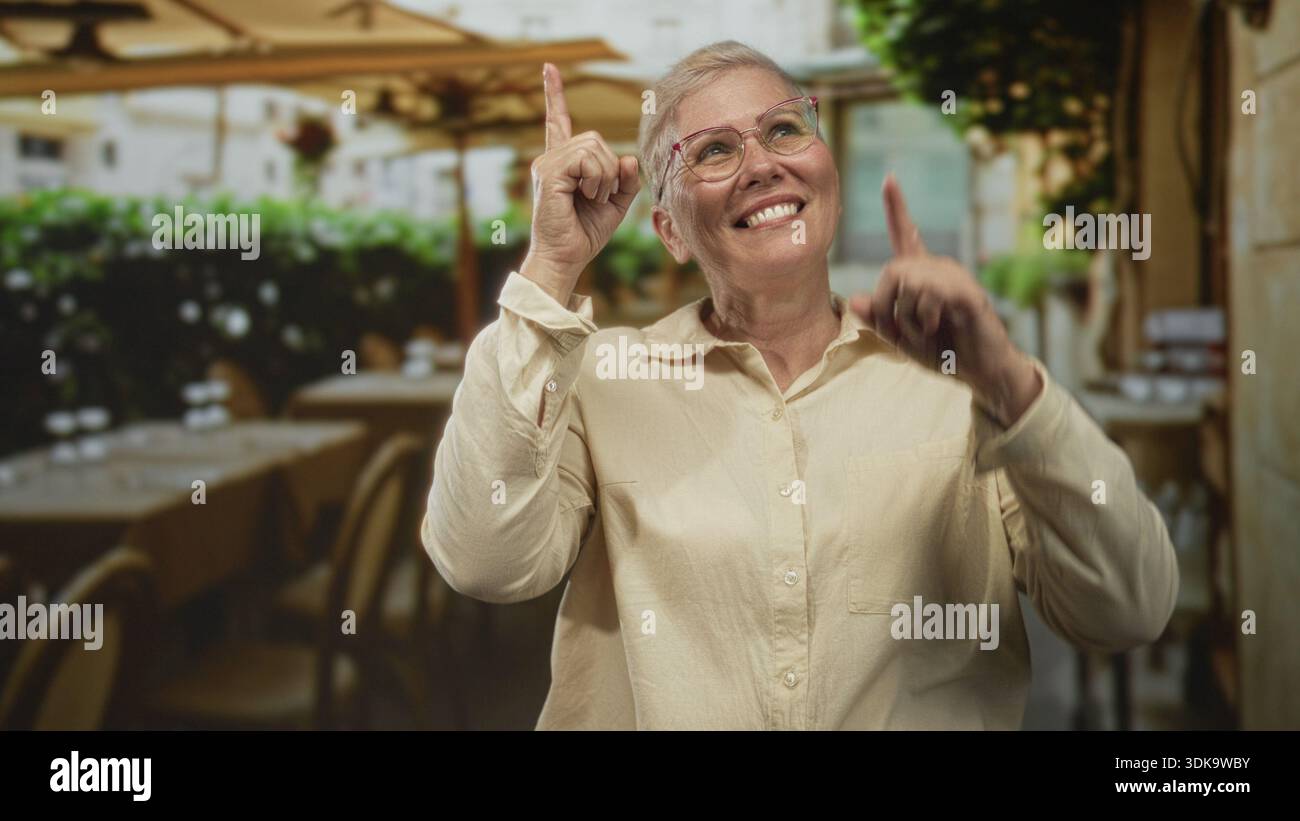 Die Frau zeigt mit den Zeigefingern auf der Straßenterrasse neben den Kaffeetischen, trägt eine Brille und lächelt; Freude feiern. Stockfoto