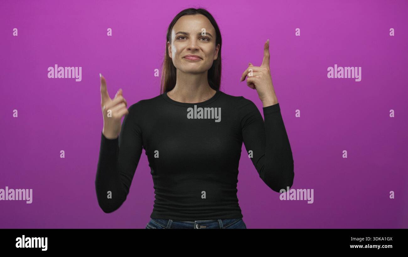 Die Frau zeigt den Zeigefinger im magentafarbenen Studio nach oben, während sie nach oben blickt und ein schwarzes Hemd und eine Jeans trägt; Neugier. Stockfoto