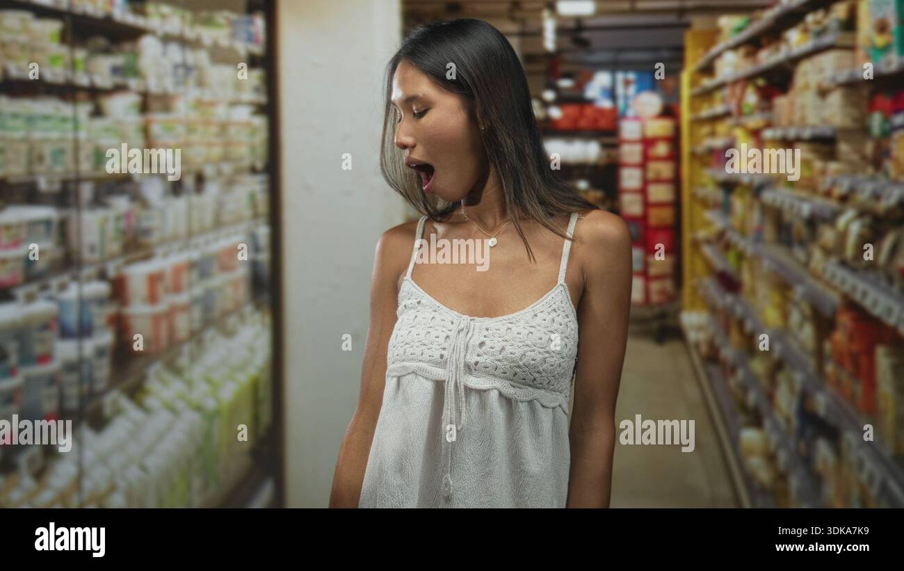 Thailändische Frau mit freiliegender Schulter, weißem Häkeltop, Kopf gedreht, Blick nach unten und Mund offen in einem Gähnen im Supermarkt; müde Besinnung. Stockfoto