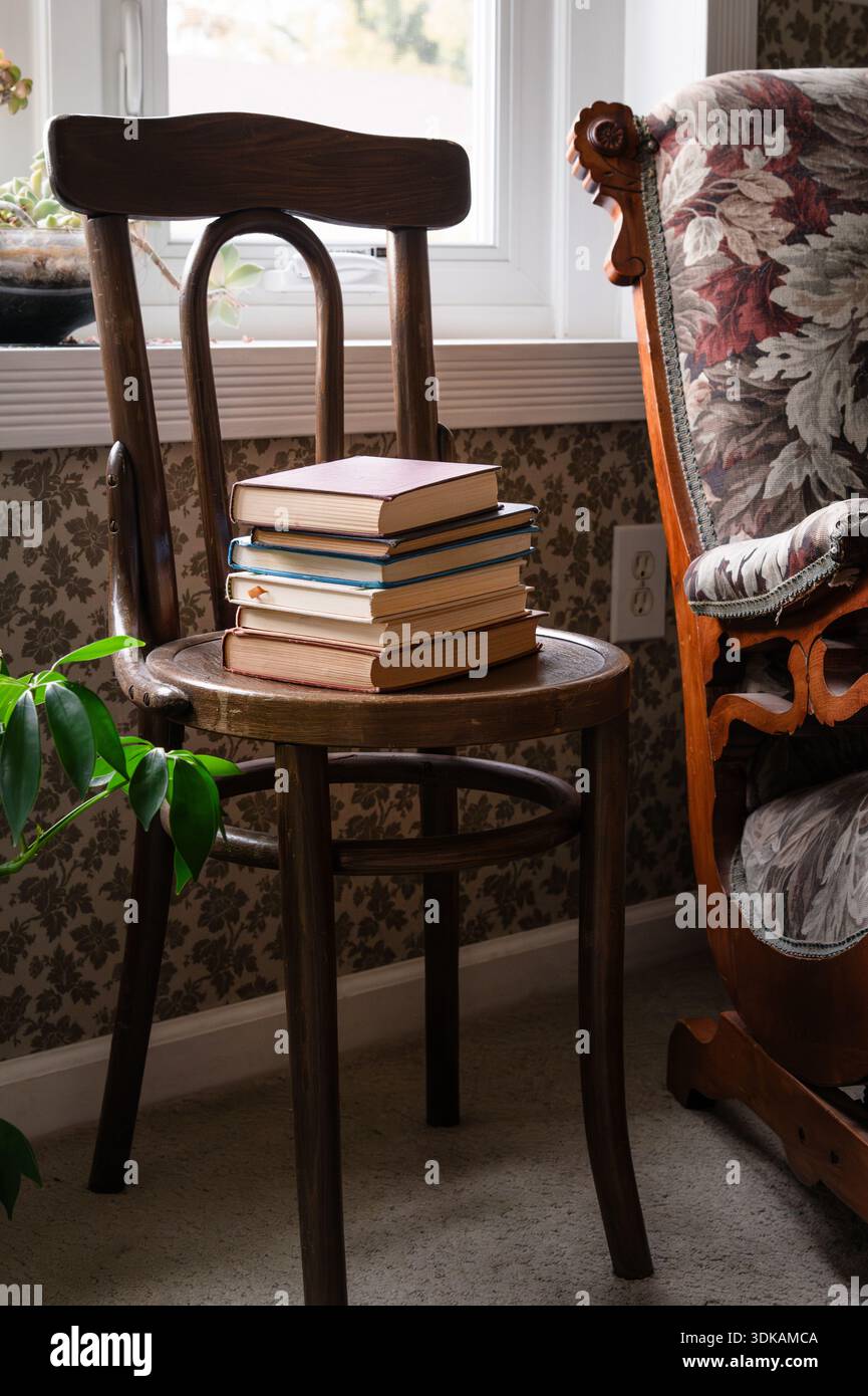Stapel alter Bücher auf einem Holzstuhl vor dem Fenster mit Pflanzen Stockfoto