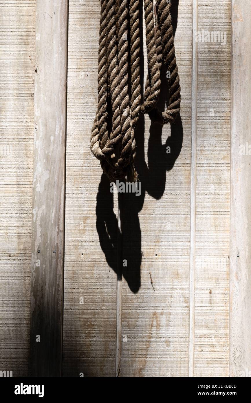 Ein gewickeltes Seil hängt an einer verwitterten Holzwand und wirft einen starken Schatten bei direktionalem Sonnenlicht. Stockfoto