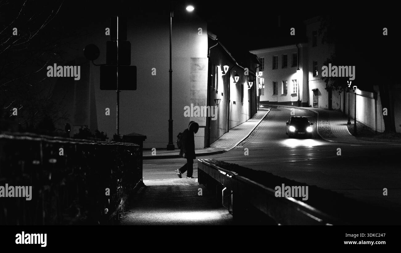 Einsame Figur, die nachts auf einer ruhigen Straße steht, von Straßenlaternen beleuchtet, mit einem sich nähernden Auto im Hintergrund. Schwarz-weiße Flosse im Kino Stockfoto
