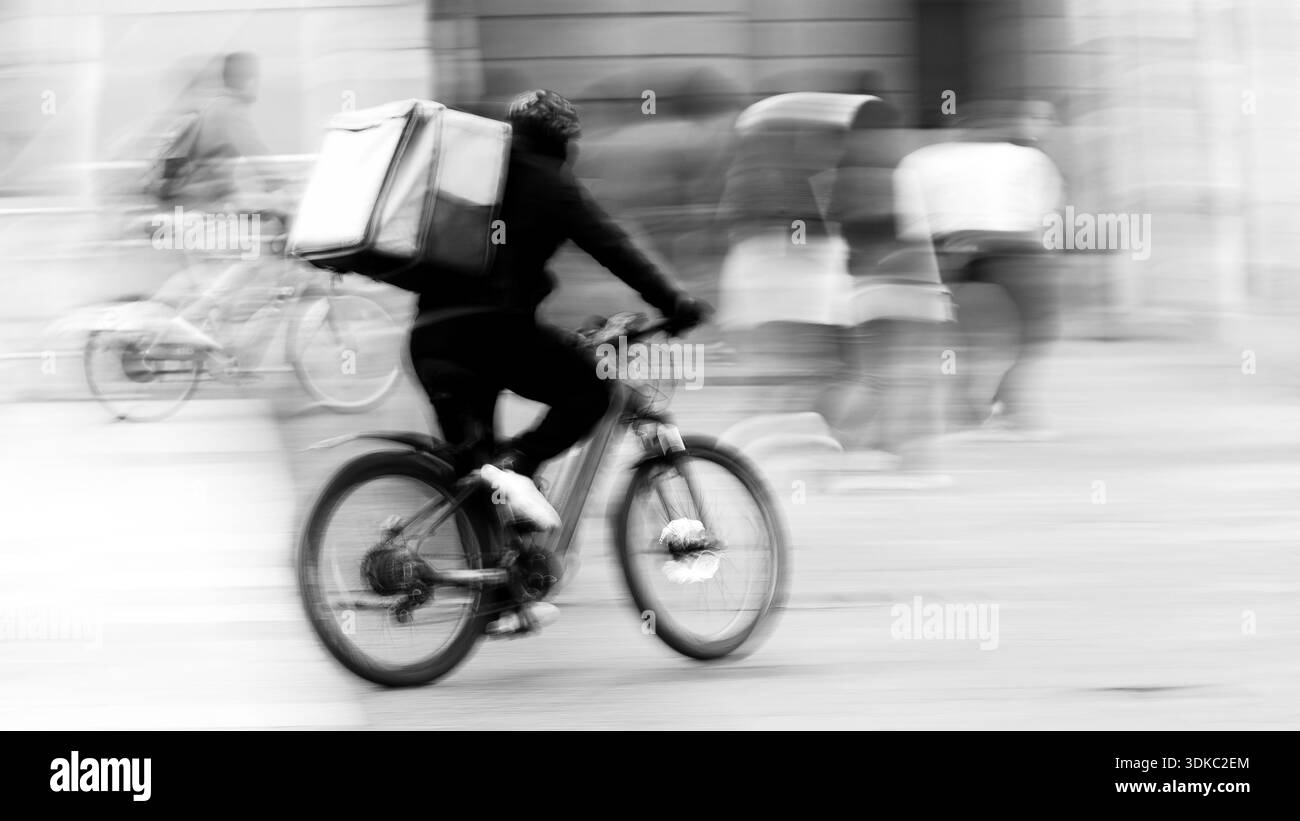 Essenskurier fährt mit dem Fahrrad durch eine geschäftige Stadtstraße mit Bewegungsunschärfe. Schwarz-weiß-urbane Szene, die Geschwindigkeit und modernen Lieferkult zeigt Stockfoto