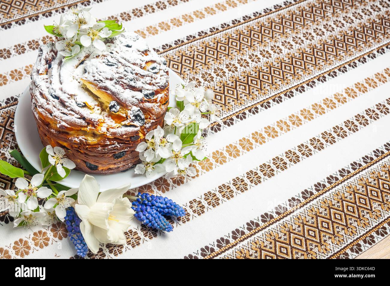 Ein wunderschön gefertigtes Osterbrot, bekannt als Kulich oder Paska, geschmückt mit zarten Blüten und einem Stäuben von Konditorzucker, sitzt auf einem Festtag Stockfoto