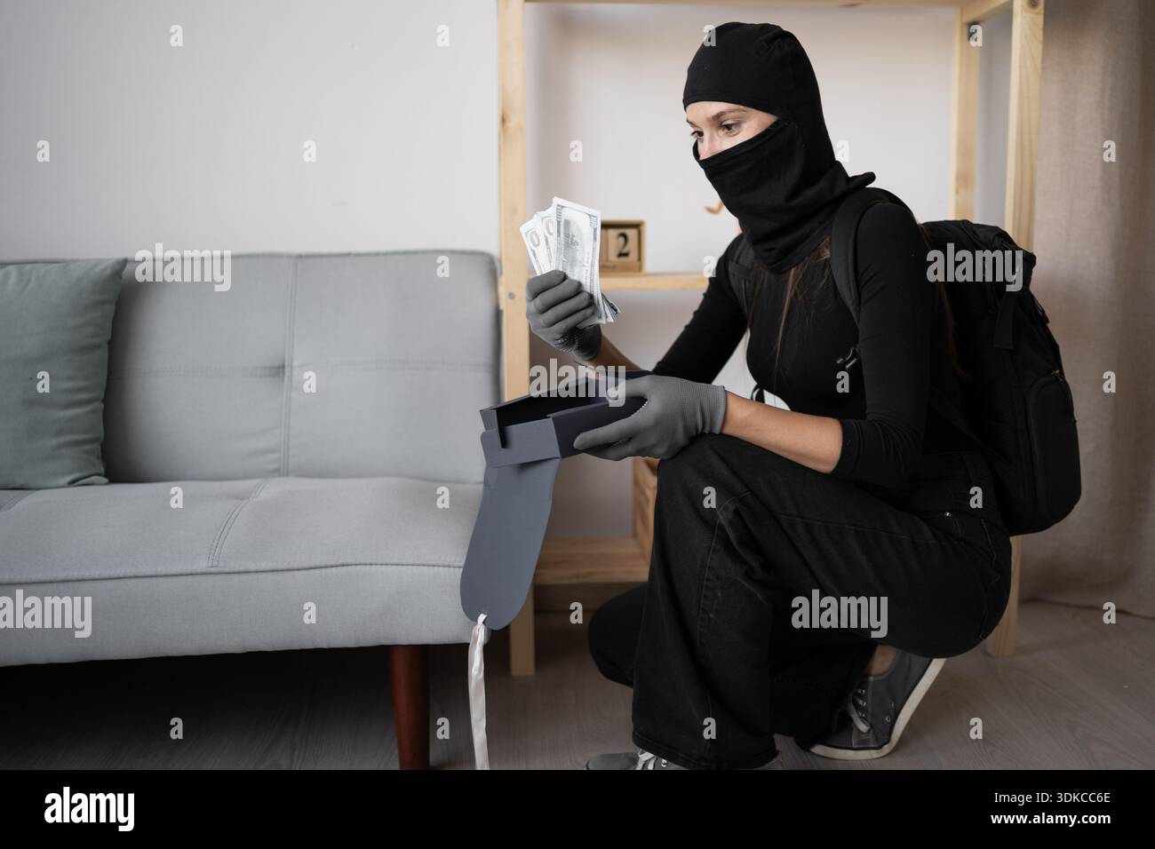 Eine Dieb in einer schwarzen Sturmhaube stiehlt zu Hause Bargeld aus einer Schreibtischschublade. Eine kriminelle Frau begeht einen Raub. Das Konzept des Hauseinbrechers Stockfoto
