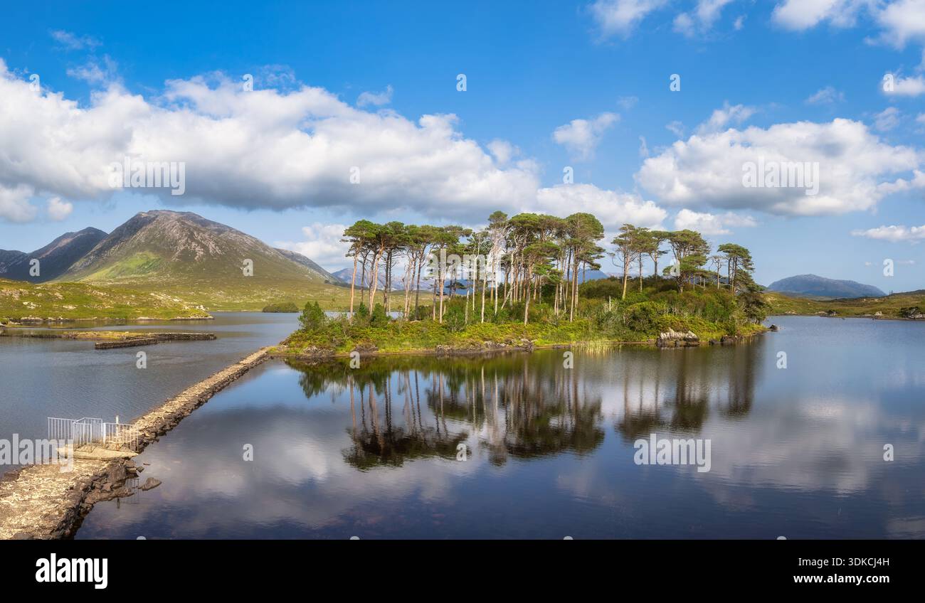 Stone Causeway führt nach Pine Island in Connemara Galway Irland. Ruhige Wasserreflexionen und windgepeitschte Bäume. Weit entfernte Berge unter dynamischen Wolken laden zu Entdeckungsreisen und Outdoor-Abenteuern ein Stockfoto