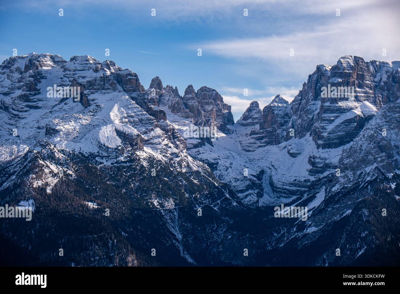 Dramatische schneebedeckte Berggipfel der Dolomiten mit geschichteten Felsformationen, die sich über einem dunklen Alpenwald unter einem klaren blauen Winterhimmel erheben, Italien. Stockfoto