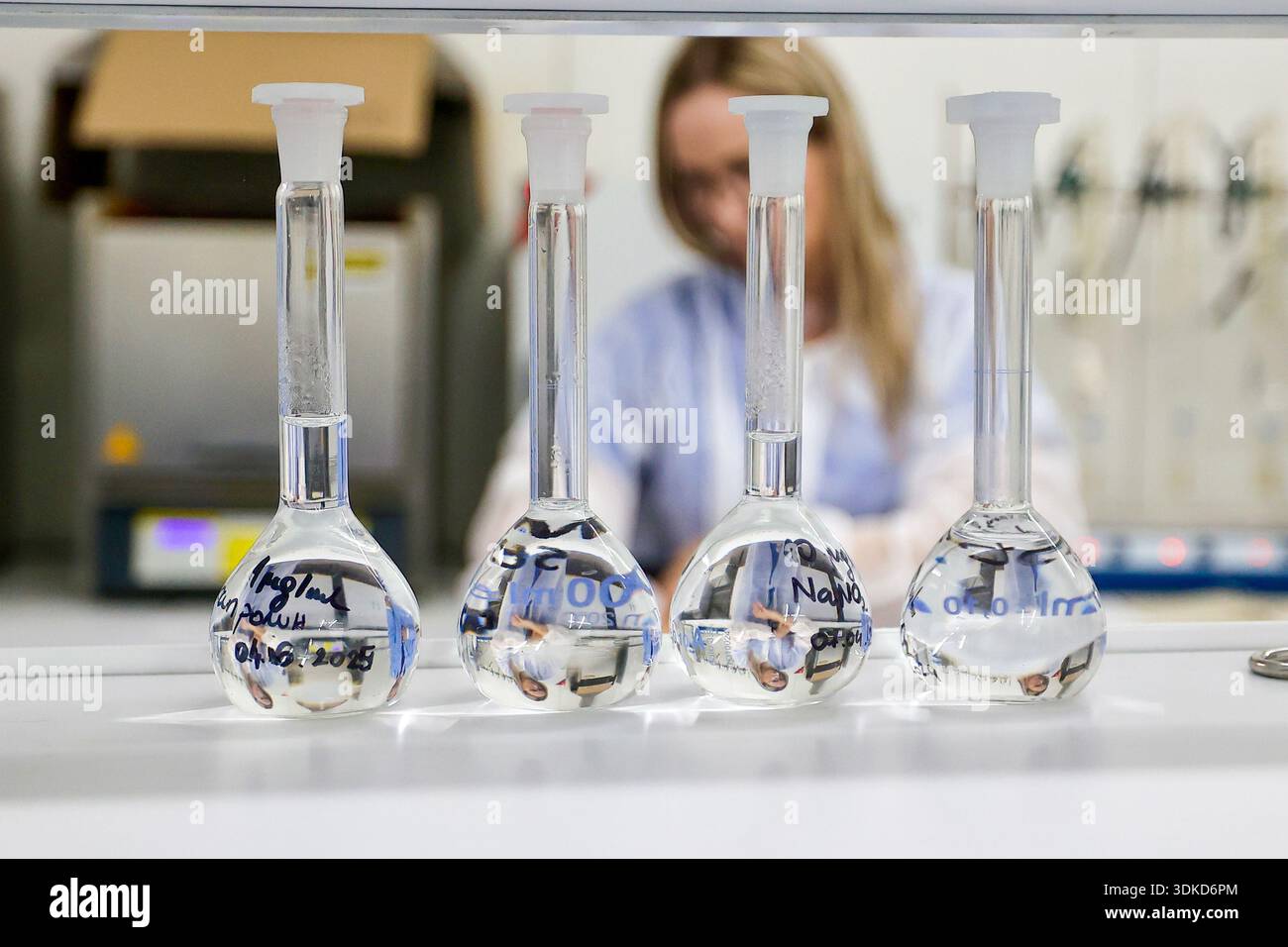 Volumetrische Laborfläschchen mit klarer Flüssigkeit auf einer Arbeitsplatte, mit einer Wissenschaftlerin im Hintergrund Stockfoto