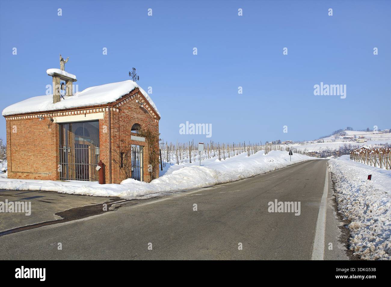 Kleine rote Kapelle am Straßenrand zwischen Weinbergen, die von weißem Schnee bedeckt sind, unter klarem blauem Winterhimmel im Piemont, Norditalien Stockfoto