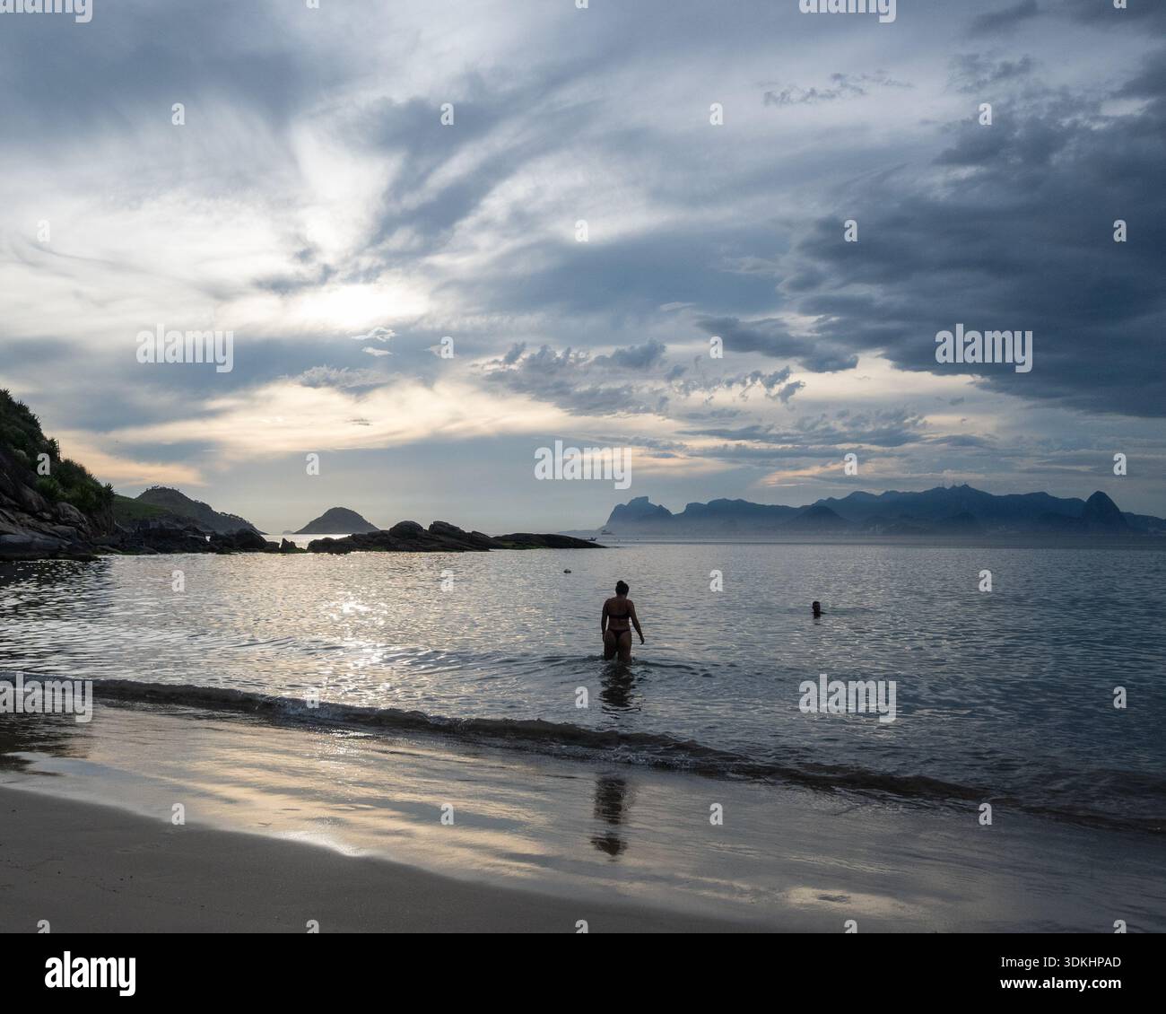 Niteroi, RJ, Brasilien - 16. Dezember 2025. Am Abend stehen Schwimmer im ruhigen Wasser am Strand von Itaipu. Dunkle Wolken, während die Sonne untergeht. Stockfoto