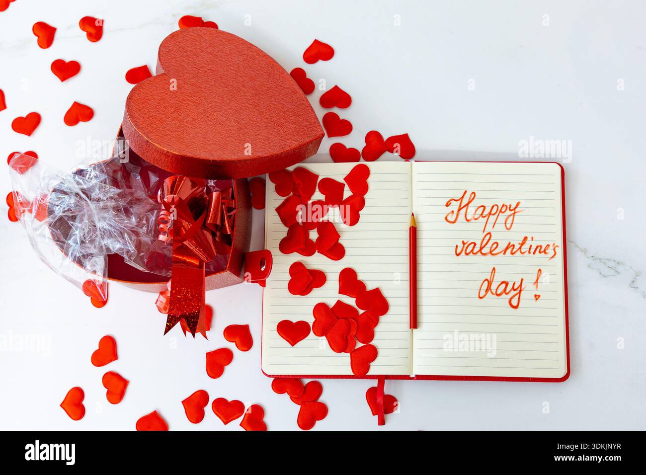 Horizontale Weitwinkelaufnahme einer offenen Geschenkbox mit rotem Herz neben einem Notizbuch mit „Happy valentine's Day!“ Text auf weißer Marmoroberfläche Stockfoto