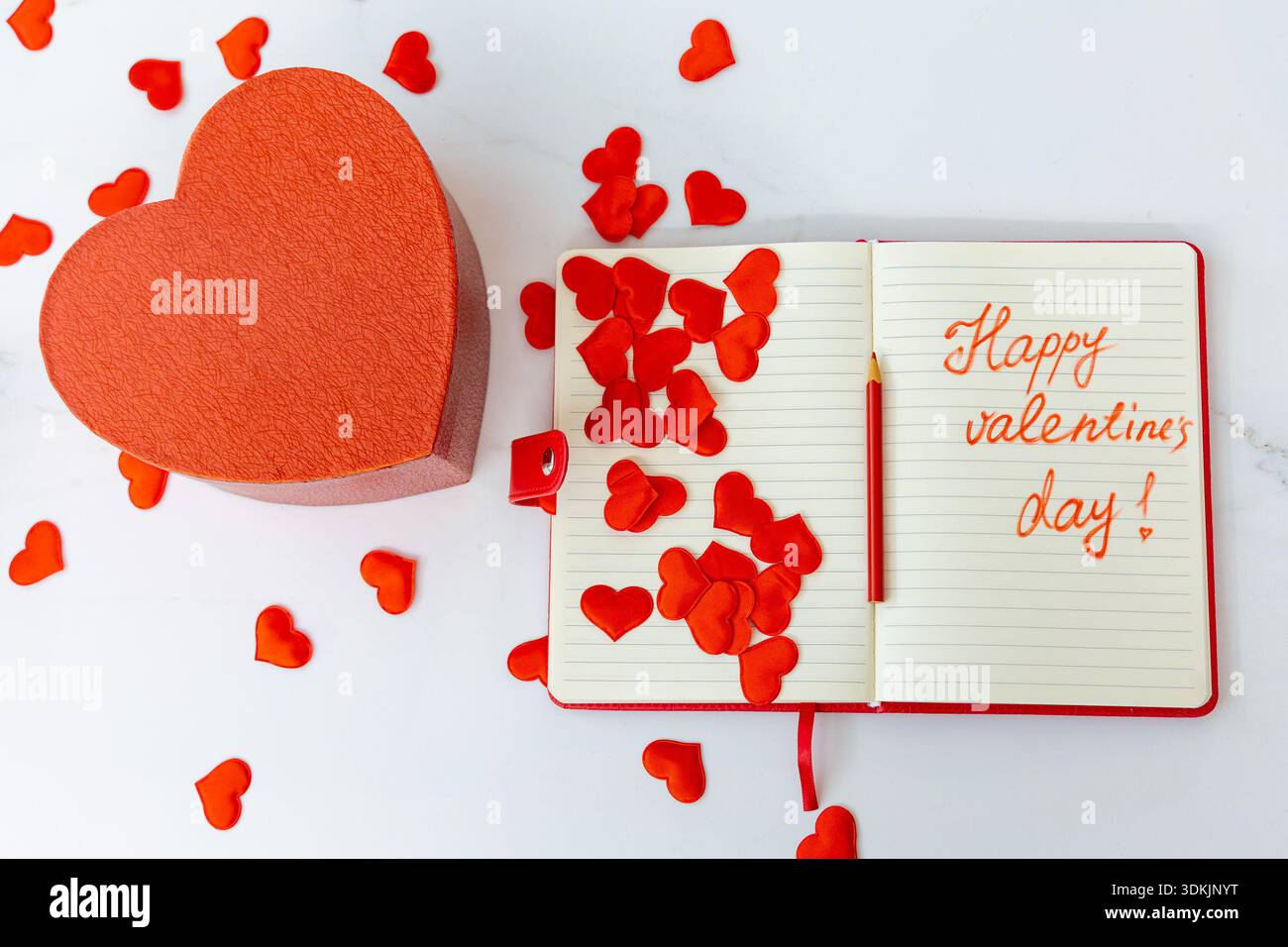 Horizontale Weitwinkelaufnahme einer offenen Geschenkbox mit rotem Herz neben einem Notizbuch mit „Happy valentine's Day!“ Text auf weißer Marmoroberfläche Stockfoto