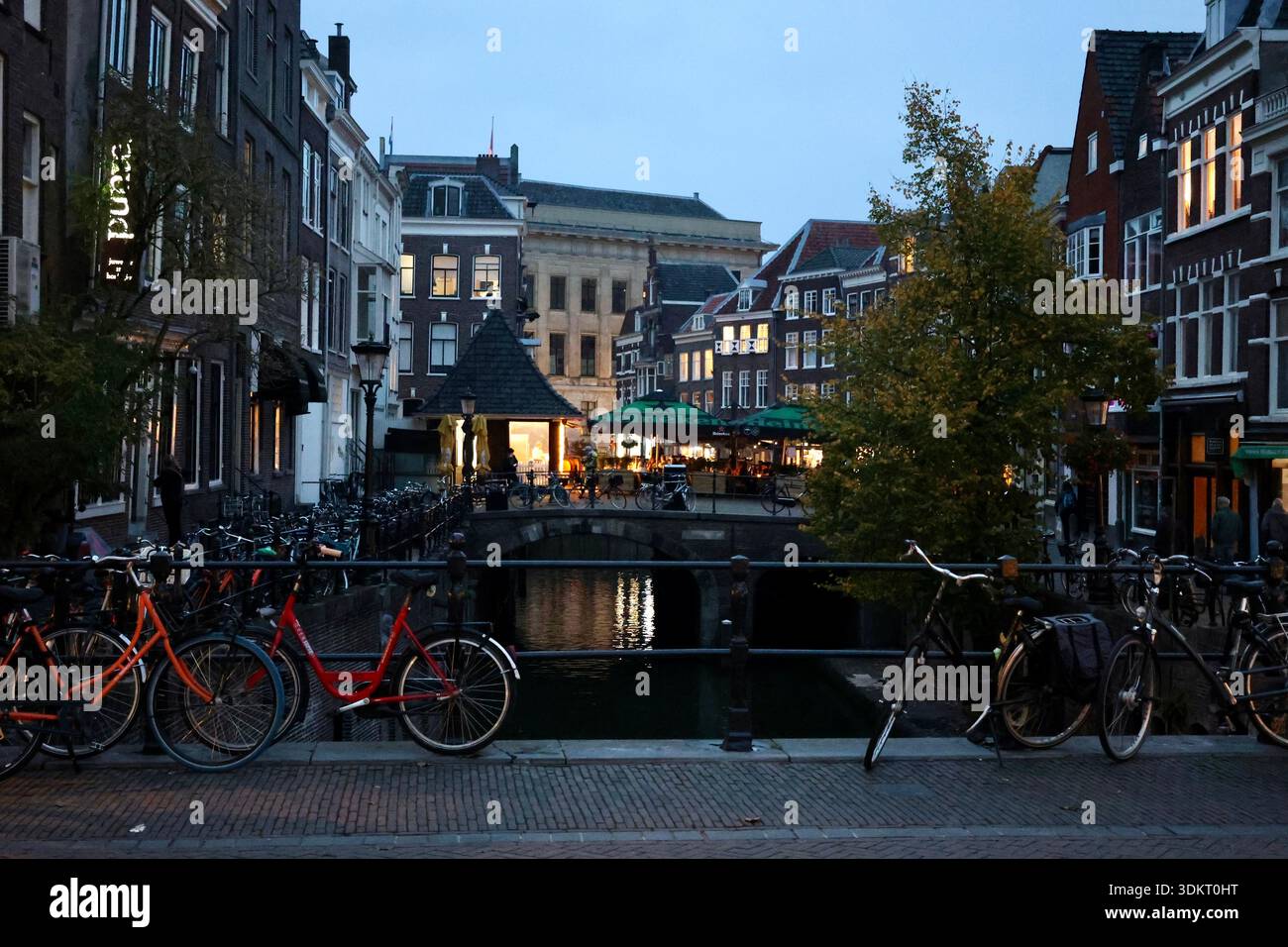 Dämmerungsblick auf einen Kanal in Amsterdam, Niederlande. Fahrräder stehen auf einer Brücke im Vordergrund, mit beleuchteten historischen Gebäuden und Bäumen im Hintergrund Stockfoto