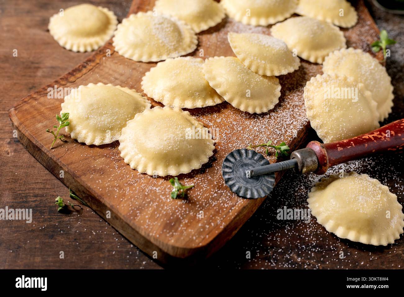 Frische hausgemachte Ravioli Nudeln auf hölzernem Schneidebrett mit Teigschneider und Kräutern. Traditionelle italienische Küche. Stockfoto