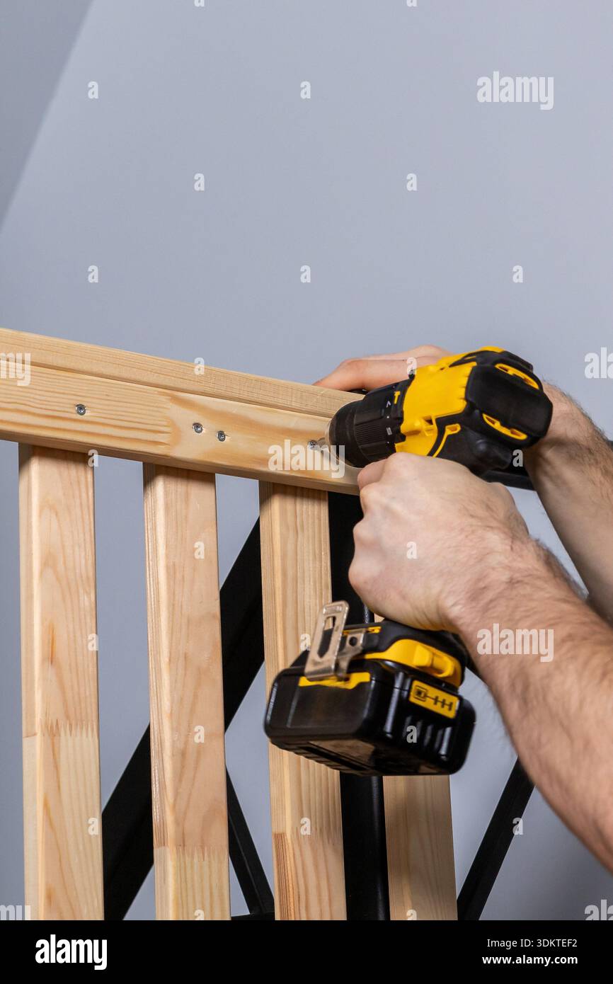 Nahaufnahme der Hände: Bedienung eines gelben und schwarzen Akku-Bohrers zum Befestigen von Schrauben in naturbelassenem Holz vor einem hellgrauen Hintergrund. Stockfoto