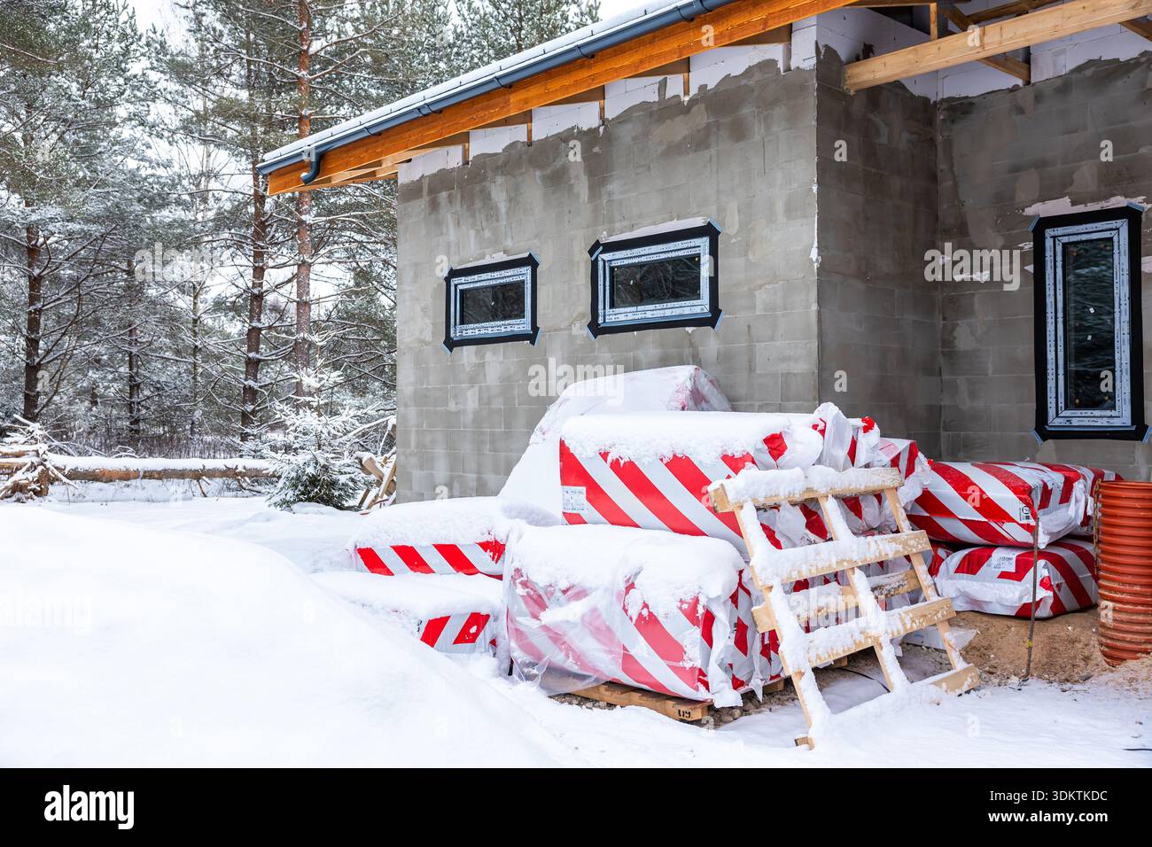 Ein im Bau befindliches Gebäude mit unvollendeten Wänden und Fenstern, umgeben von schneebedeckten Bäumen, Materialien und orangen Rohren. Stockfoto