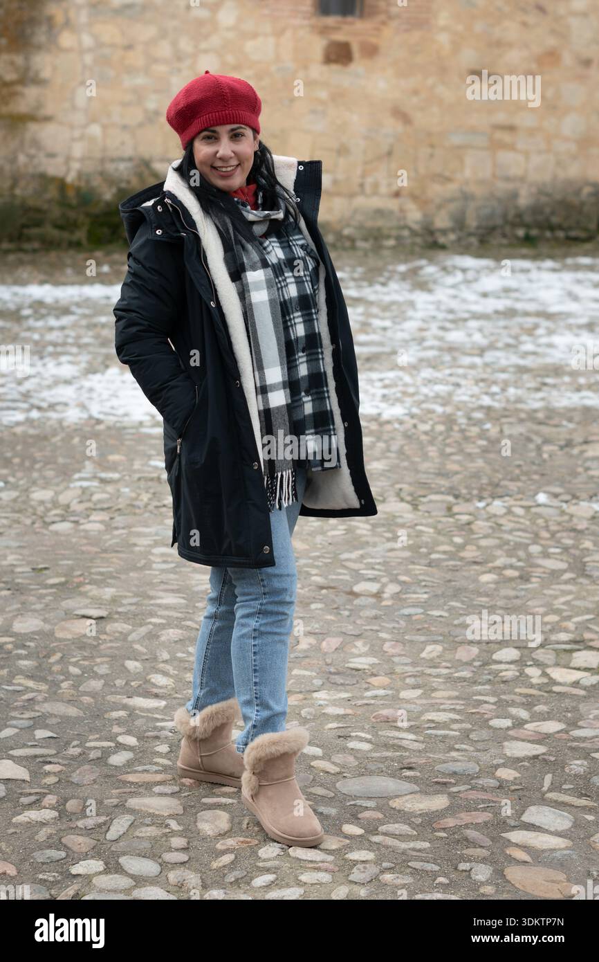 Junge Erwachsene Frau mit Wintermantel, roter Baskenmütze, kariertem Schal, Jeans und Stiefeln, draußen stehen Stockfoto