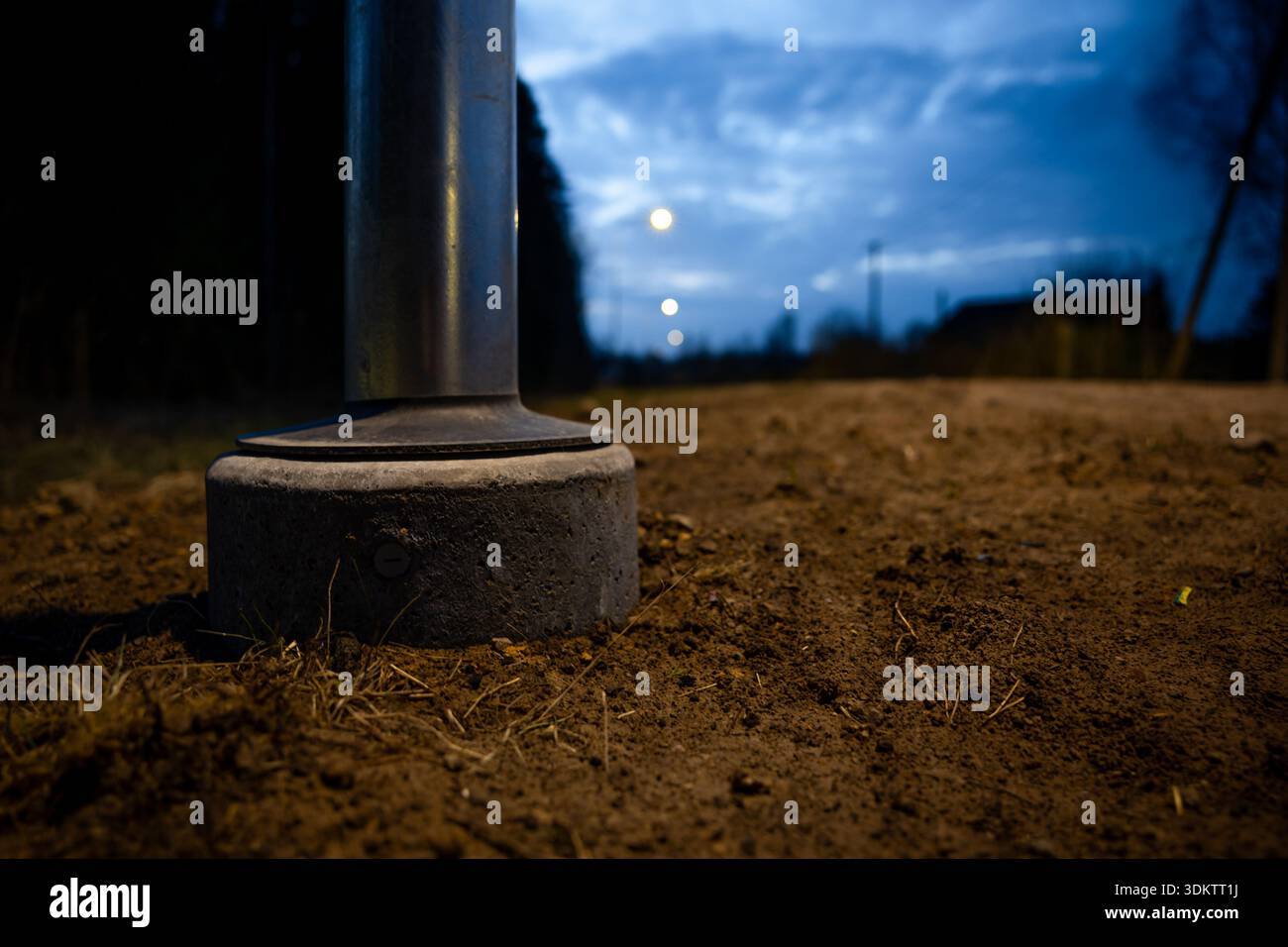 Niedriger Winkel auf die Basis eines Metallpfahls auf einem unbefestigten Weg, mit Betonfundament, schwachen Straßenlaternen, Bäumen und einem teilweise bewölkten blauen Himmel bei d Stockfoto