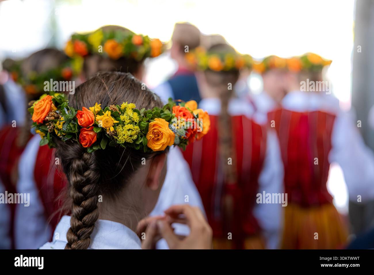 Eine Gruppe von Kindern in lettischen Nationalkostümen mit roten und weißen Streifen. Blumenkronen aus orangen und gelben Blüten zieren ihre Köpfe. Stockfoto