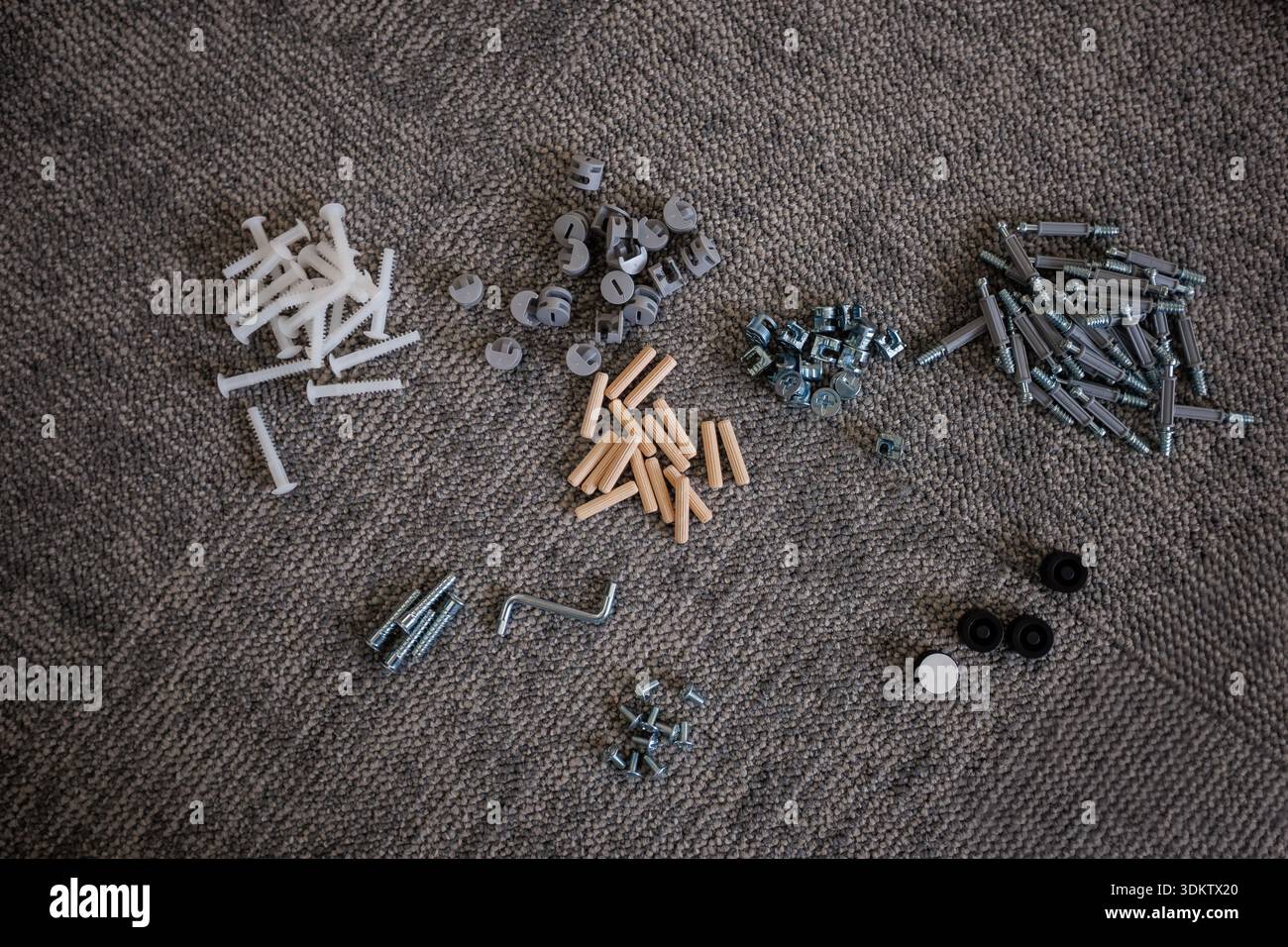 Ordentlich angeordnete Hardwarekomponenten einschließlich Schrauben, Muttern, Bolzen, Dübel, Stifte, Kappen, Räder und ein Sechskantschlüssel auf einer strukturierten grauen Oberfläche. Stockfoto