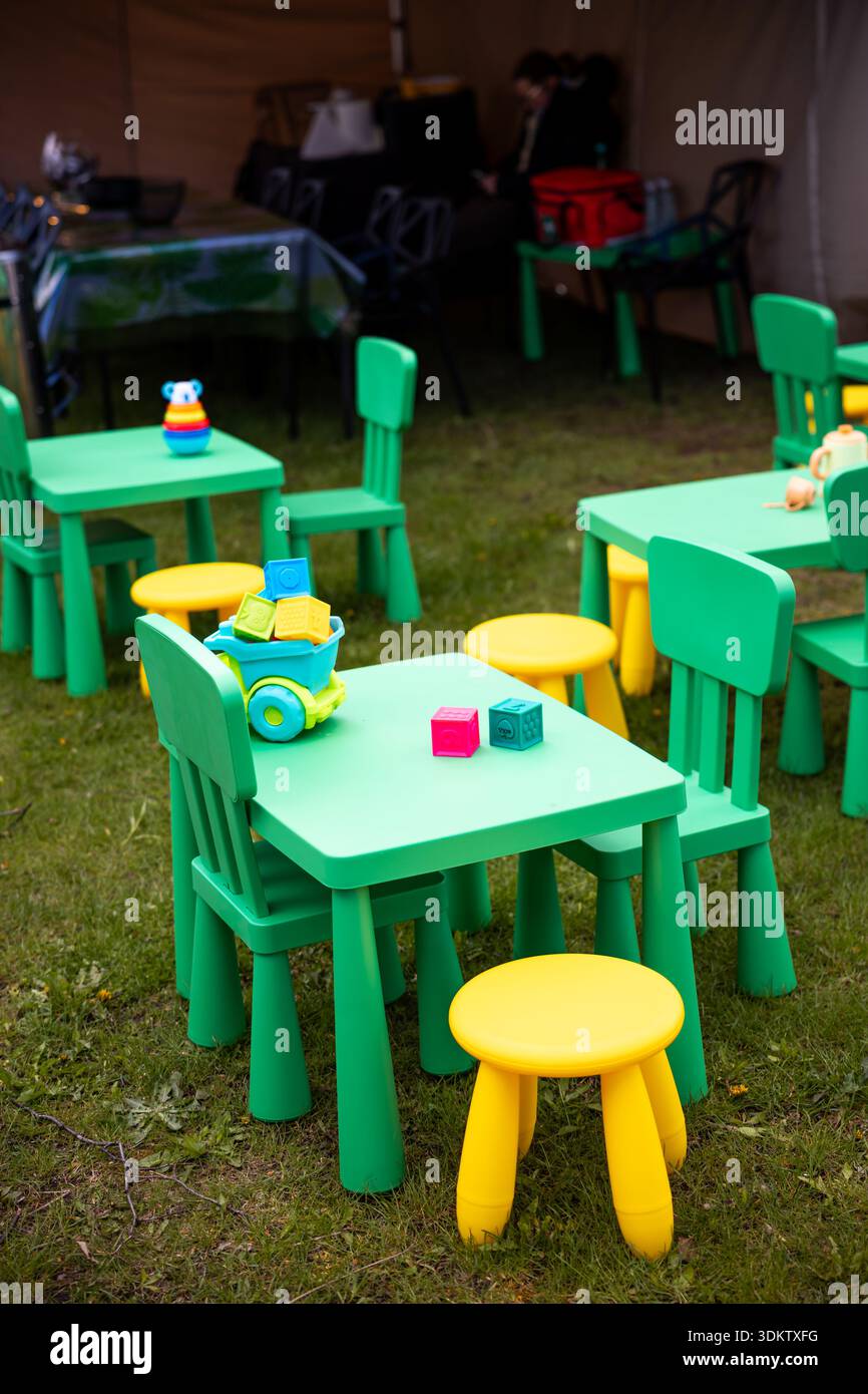 Grüne Tische, gelbe Hocker und Spielzeuge wie Blöcke und eine Spielzeugbahn befinden sich auf Gras unter einem Zelt. Im schattigen Bereich befinden sich zusätzliche Sitzplätze. Stockfoto