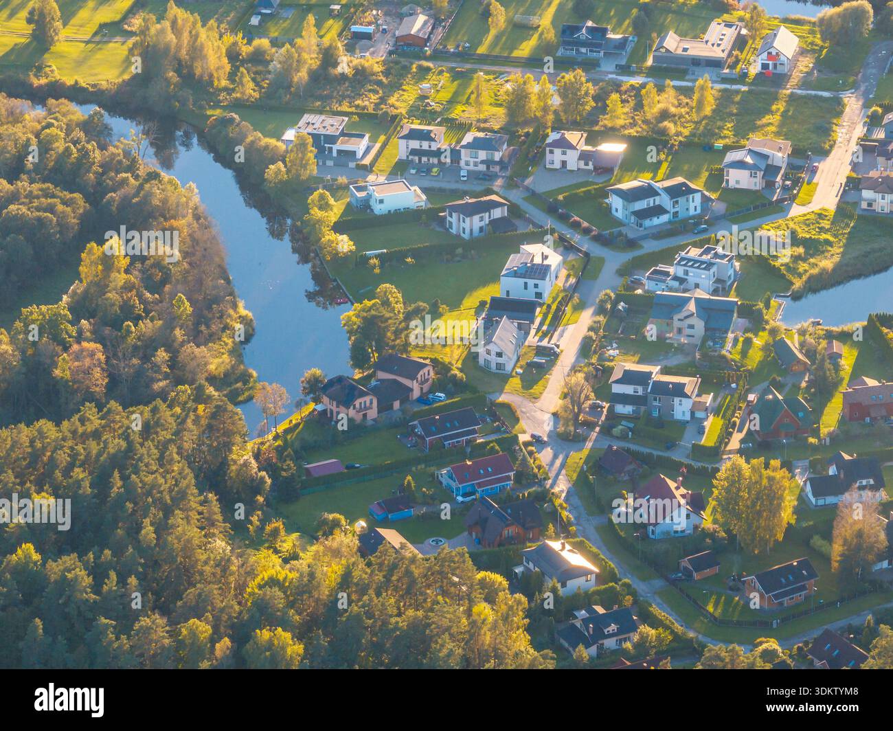 Aus der Vogelperspektive eines Wohnviertels mit gewundenen Straßen, einem reflektierenden Wasserweg, offenen Grünflächen und Baumgruppen in sanftem Sonnenlicht. Stockfoto