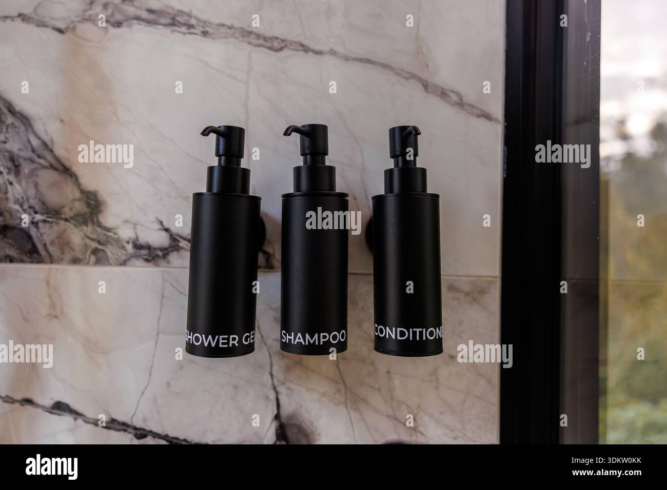 Drei schwarze Pumpflaschen mit der Aufschrift Duschgel, Shampoo und Conditioner sind an einer Wand mit grauen und weißen Adern montiert. Auf t ist eine Glasscheibe sichtbar Stockfoto