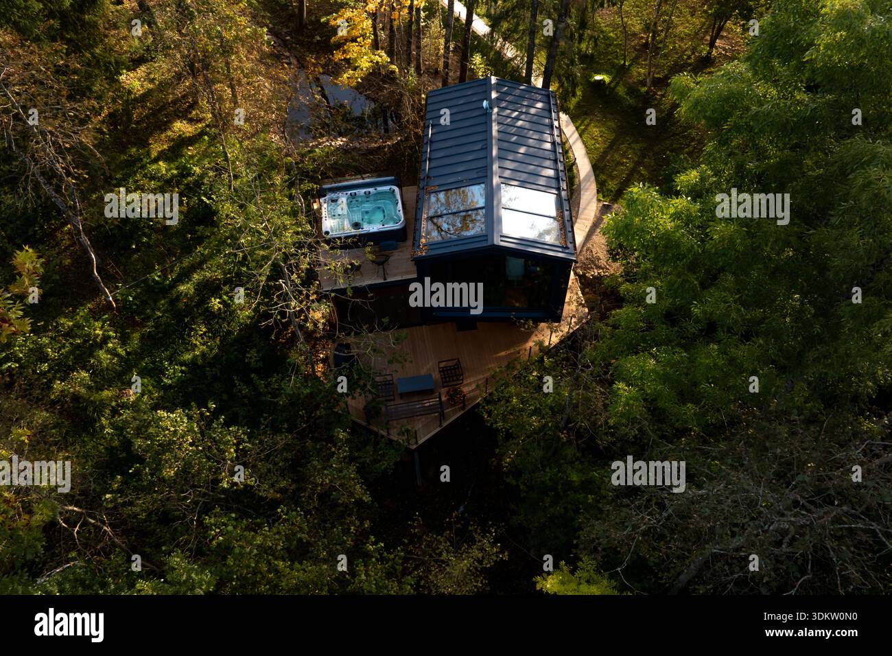 Eine moderne Hütte mit großen Glasfenstern, eckigem Dach und Holzterrasse mit Sitzgelegenheiten und Whirlpool, umgeben von Bäumen und Sonnenlicht, das nach außen filtert Stockfoto