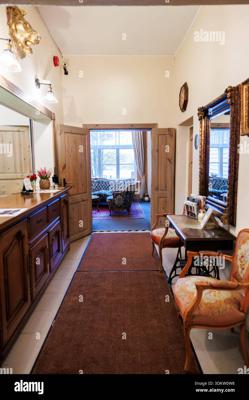 Hotelfoyer im Vintage-Stil mit Rezeption, vergoldetem Spiegel und Blick auf den Salon. Schreibtisch aus poliertem Holz, Löwenkopf Wandleuchte, Uhr, antike Stühle, roter Teppich, weiche Lampen, Stockfoto
