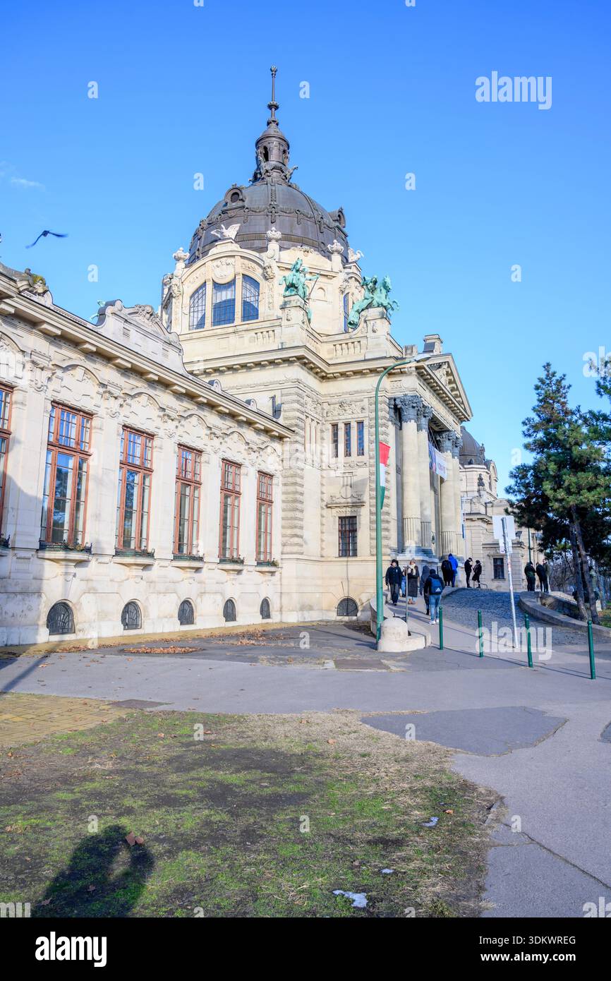 Das Szechenyi Thermalbad von außen zeigt seine kunstvolle neobarocke Architektur mit einer Kuppel und Statuen unter einem klaren blauen Himmel, wie es die Besucher tun Stockfoto