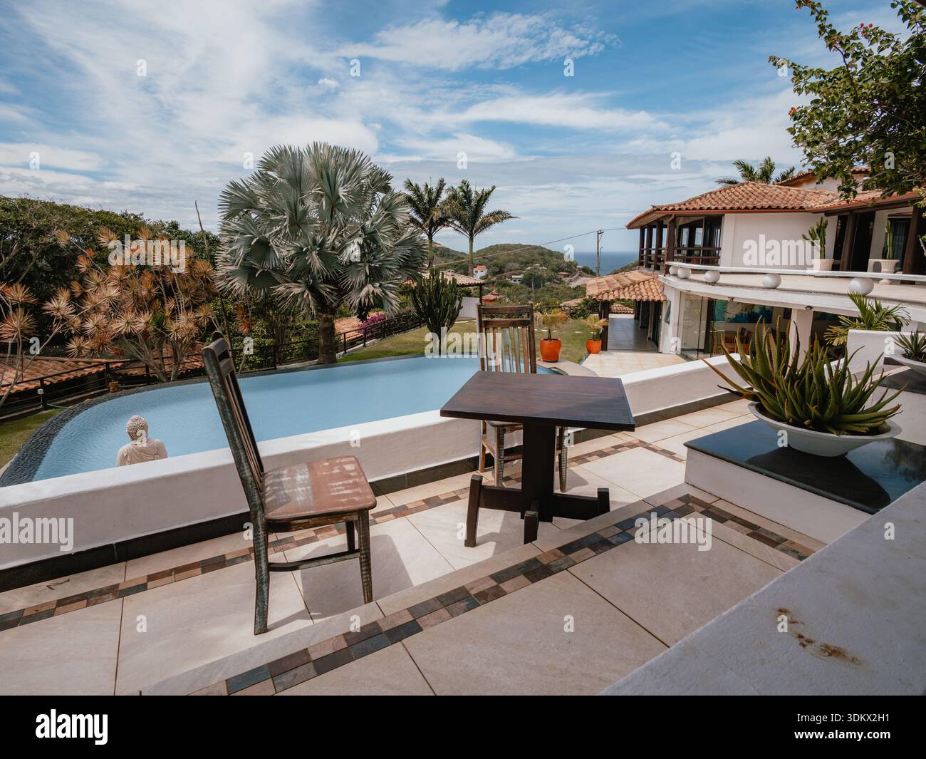 Outdoor-Szene mit einer möblierten Terrasse vor einem Swimmingpool und einem Villa-ähnlichen Haus, mit Vegetation und teilweise bewölktem Himmel. Perfektes Bild für Profis Stockfoto