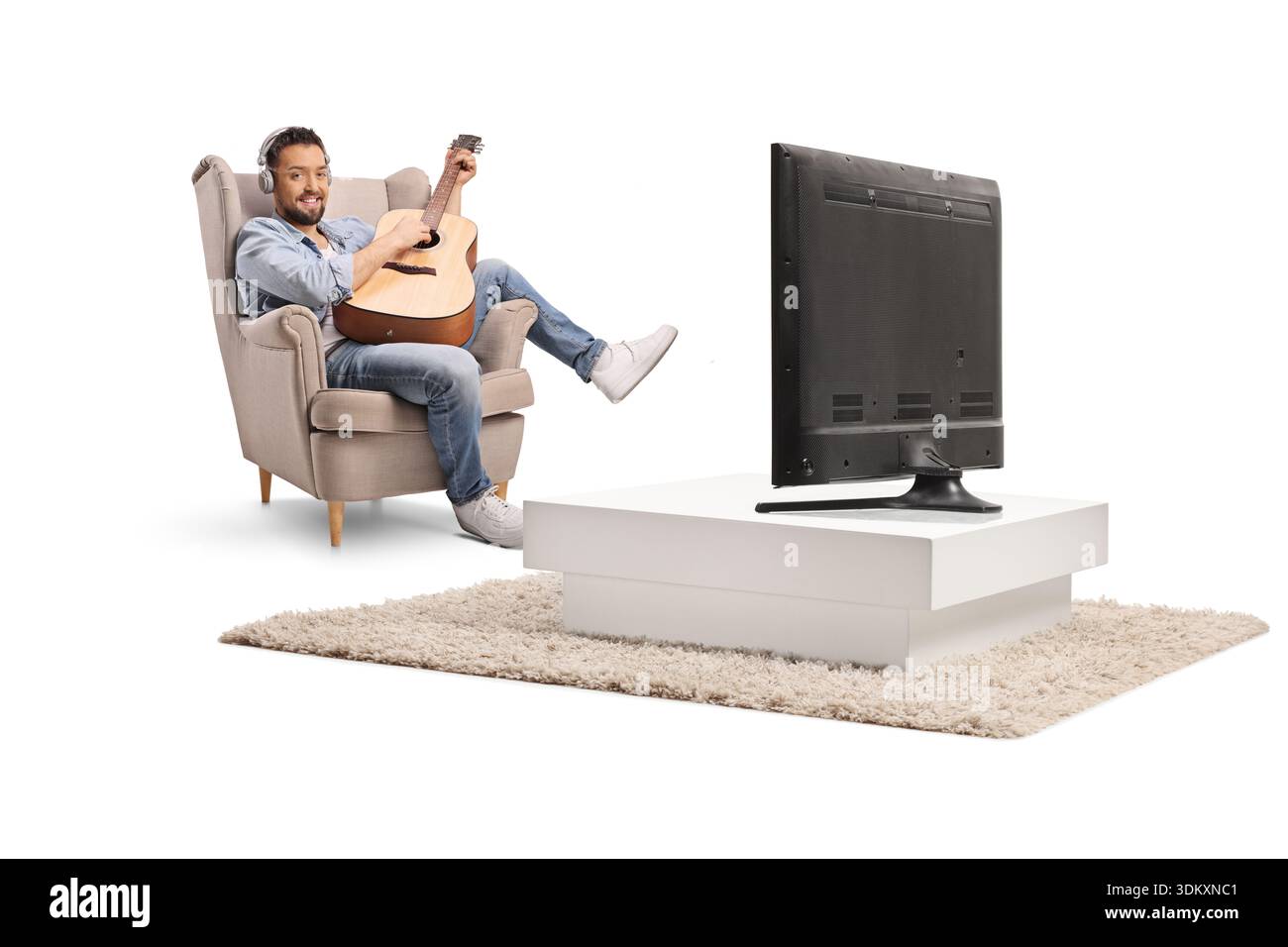Junger Mann in einem Sessel vor dem fernseher, der eine Akustikgitarre spielt, isoliert auf grauem Hintergrund Stockfoto