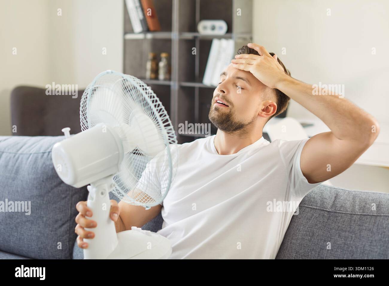 Erschöpfter Mann, der sich mit Ventilator kühlt, sitzt auf der Couch im Wohnzimmer Stockfoto
