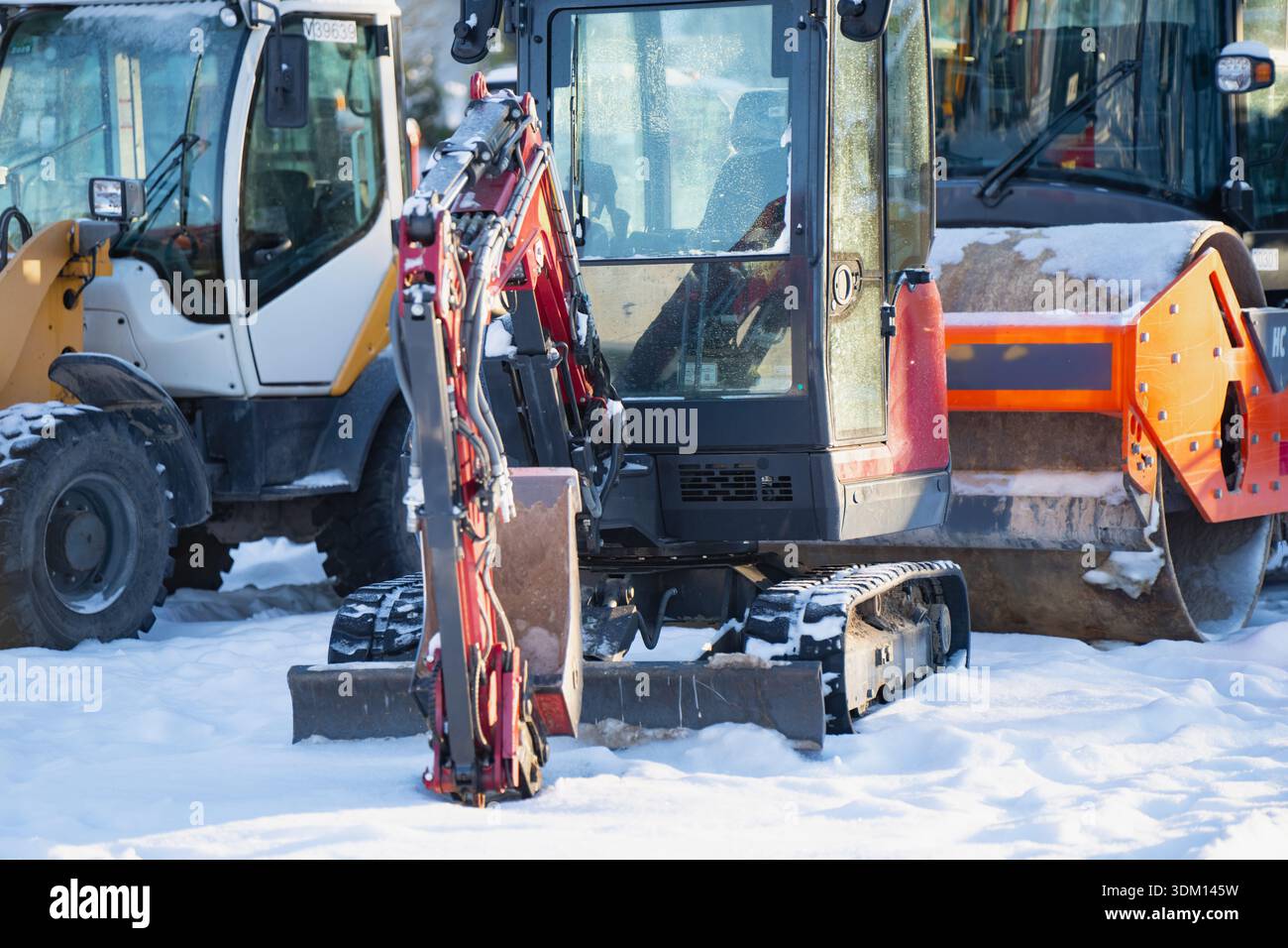 Kompakter Bagger auf Schnee im Maschinenhof geparkt, Vorderansicht mit Kabinenglas, rotem Hydraulikarm und Gummiketten, mit größeren Maschinen und Schaufel Stockfoto