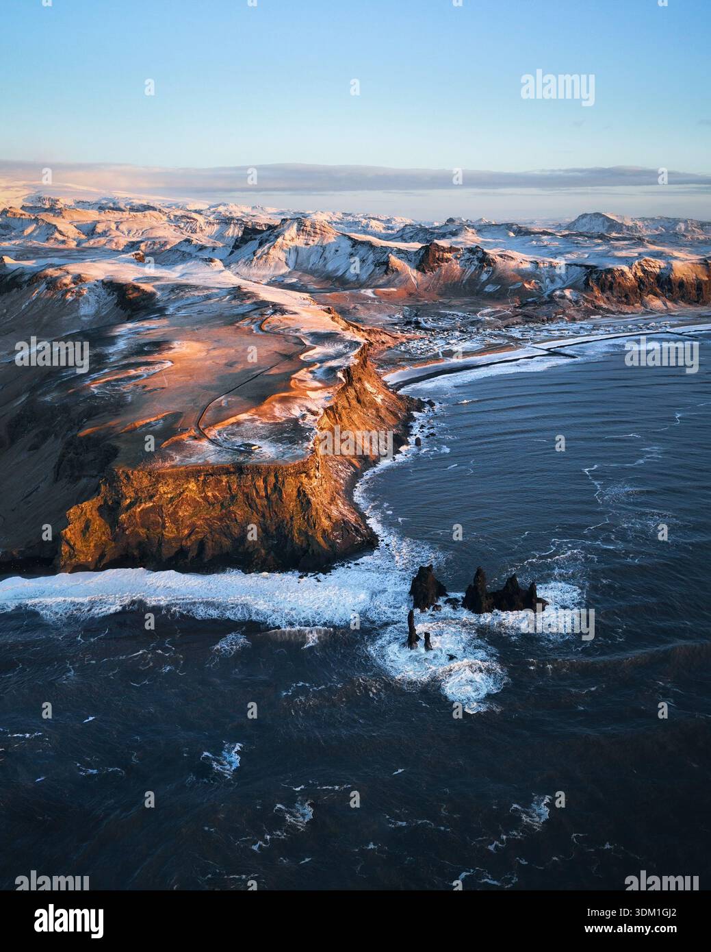 Blick aus der Vogelperspektive auf die steilen Klippen, die auf das turbulente Meer treffen, das goldene Licht, das die Kanten der schneebedeckten Gipfel küsst, ein Tanz aus Feuer und Eis, Island. Stockfoto