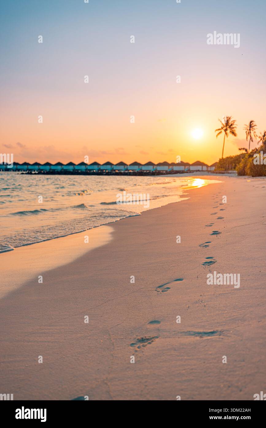 Atemberaubender tropischer Strand Sonnenuntergang Fußabdrücke Sand führende Wasservillen warmer goldener Himmel vertikal einsam romantische Dämmerung ruhiger traumhafter Hintergrund Stockfoto