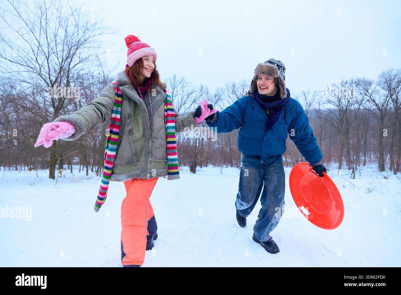 Ein junges Paar, das im verschneiten Park läuft und Hände mit dem Schlitten hält. Stockfoto