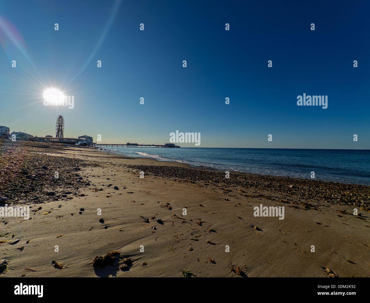 Sonnendurchflutete Worthing-Küste mit hoch aufragendem Riesenrad und Pier vor Kieselstrand und ruhigem Englischen Kanal, blauer Himmel über dem Meer. - Smartphone-aufgenommenes Stockfoto