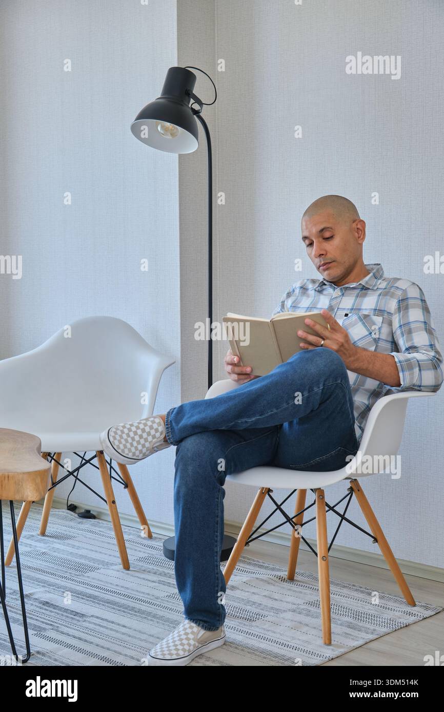 Mann liest ein Buch, während er auf einem modernen Stuhl in einem minimalistischen Wohnzimmer sitzt Stockfoto