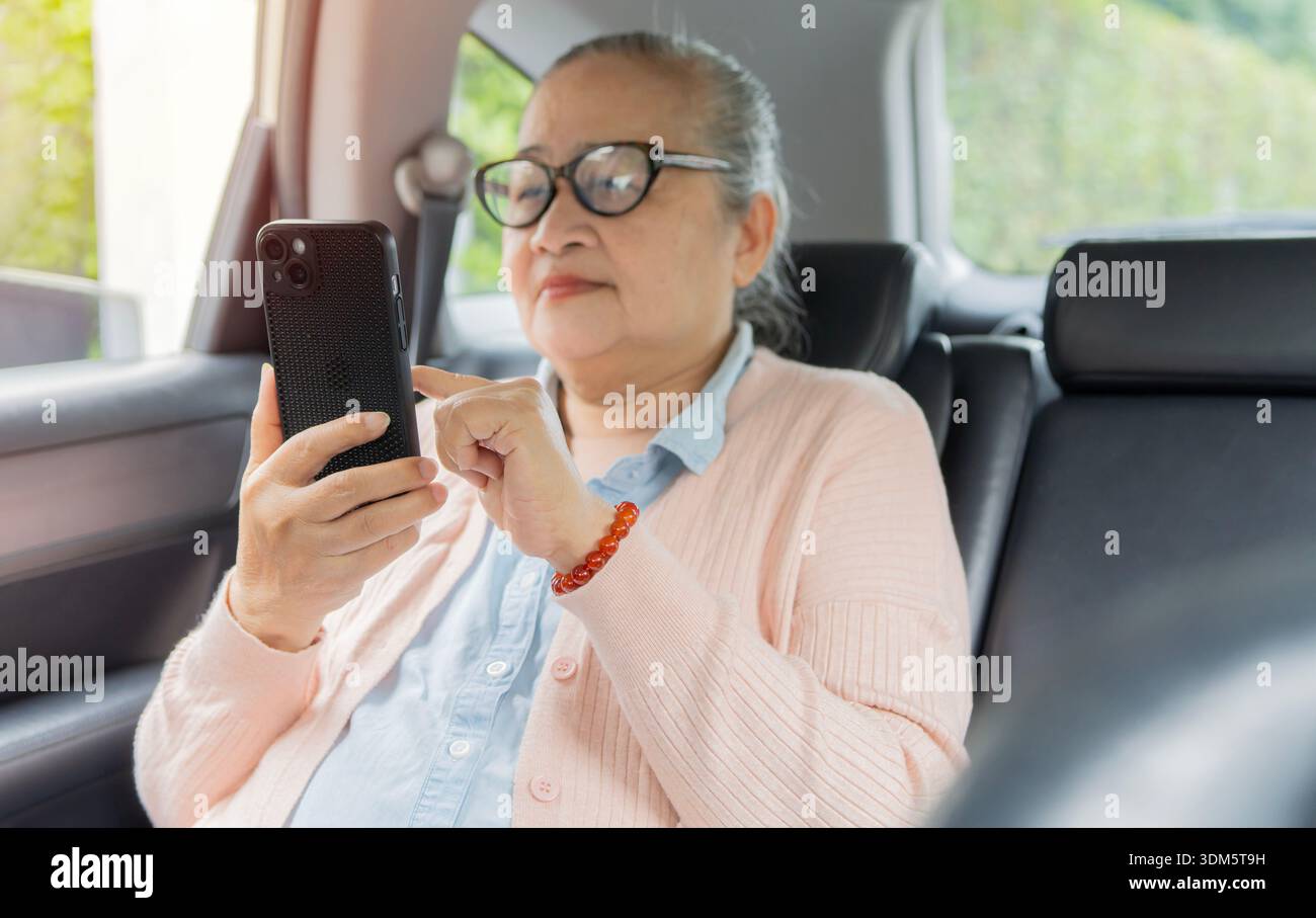 Ältere asiatische Frau, die ihr Smartphone benutzt, während sie im modernen Auto auf dem Rücksitz sitzt. Lebensstil-Konzept im Ruhestand. Stockfoto