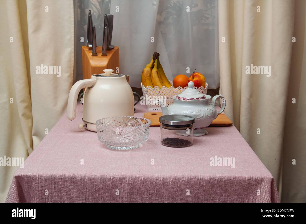 Auf einer rosafarbenen Tischdecke befinden sich eine Sammlung von Küchenartikeln, darunter eine Teekanne, ein Wasserkocher, eine Obstschale und ein Messerblock Stockfoto