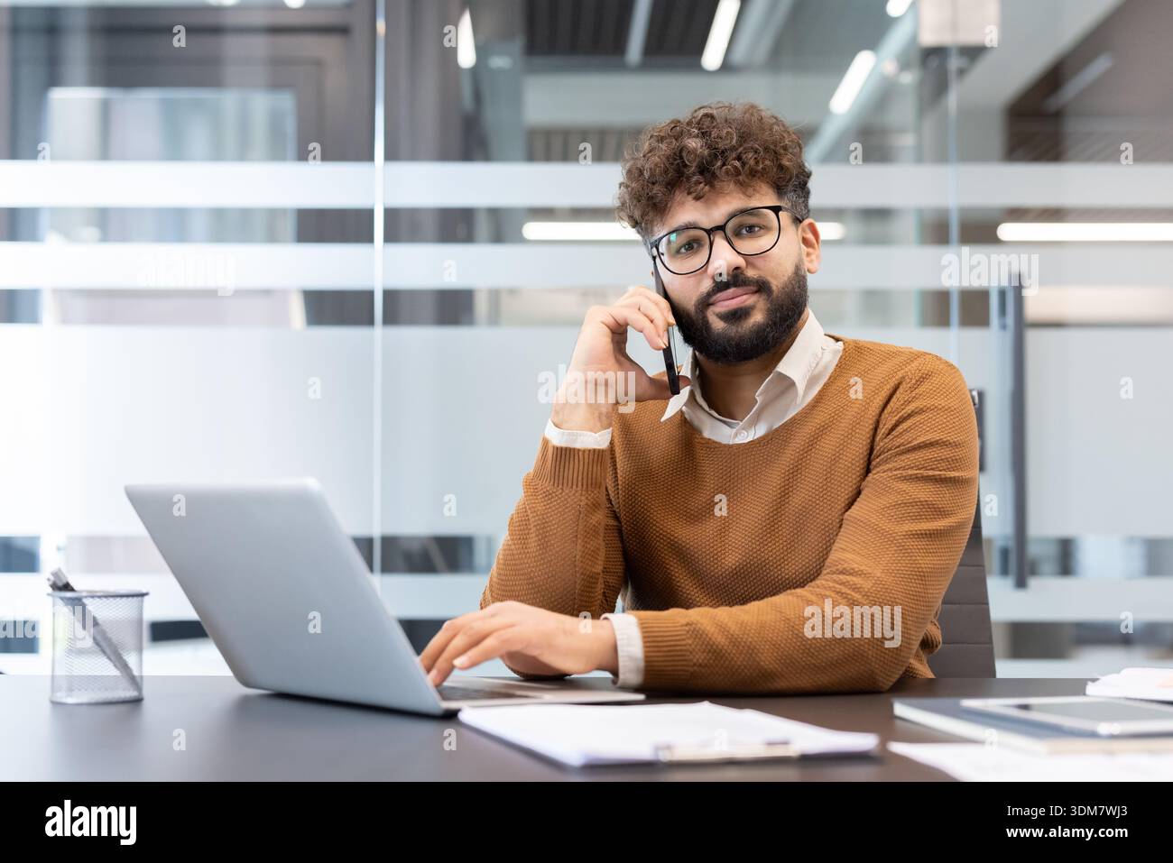 Junger Geschäftsmann mit lockigem Haar und Brille, der über sein Mobiltelefon kommuniziert, während er an einem modernen Arbeitsplatz an einem Laptop arbeitet und Geschäftsabläufe verwaltet Stockfoto