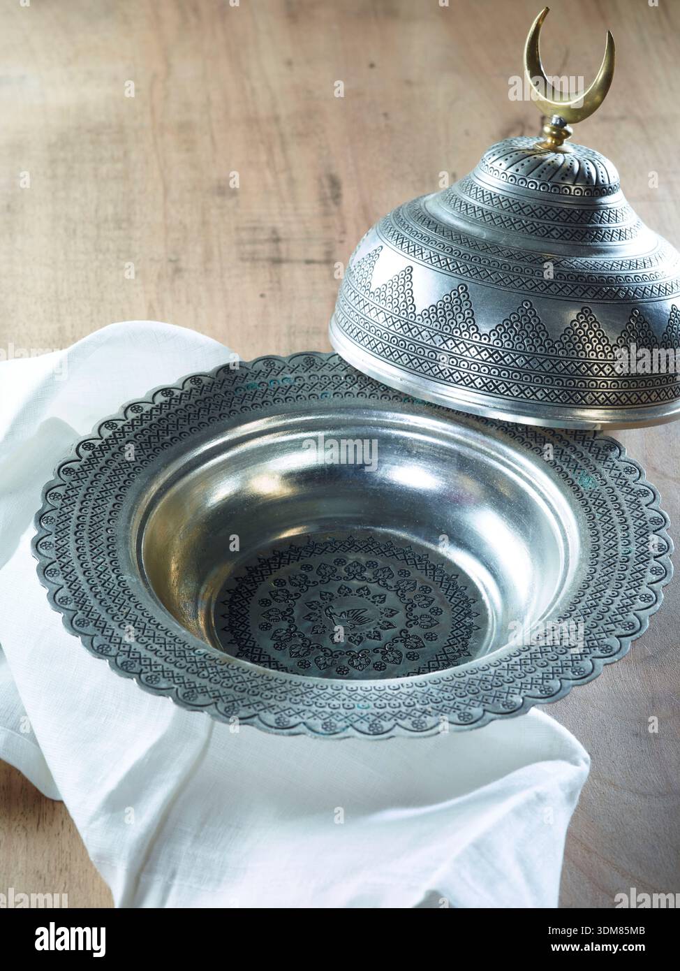 Traditionelle, gravierte Metallschale mit halbmondförmigem Deckel auf rustikalem Holztisch, Ramadan-Essen Stockfoto
