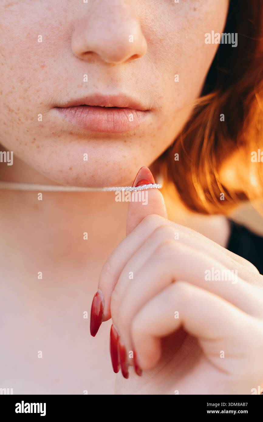 Eine sanfte und intime Nahaufnahme einer jungen rothaarigen Frau, die eine zarte Diamantkette in der Nähe ihrer Lippen hält. Ihre natürlichen Sommersprossen, sanften rosa Lippen und Stockfoto
