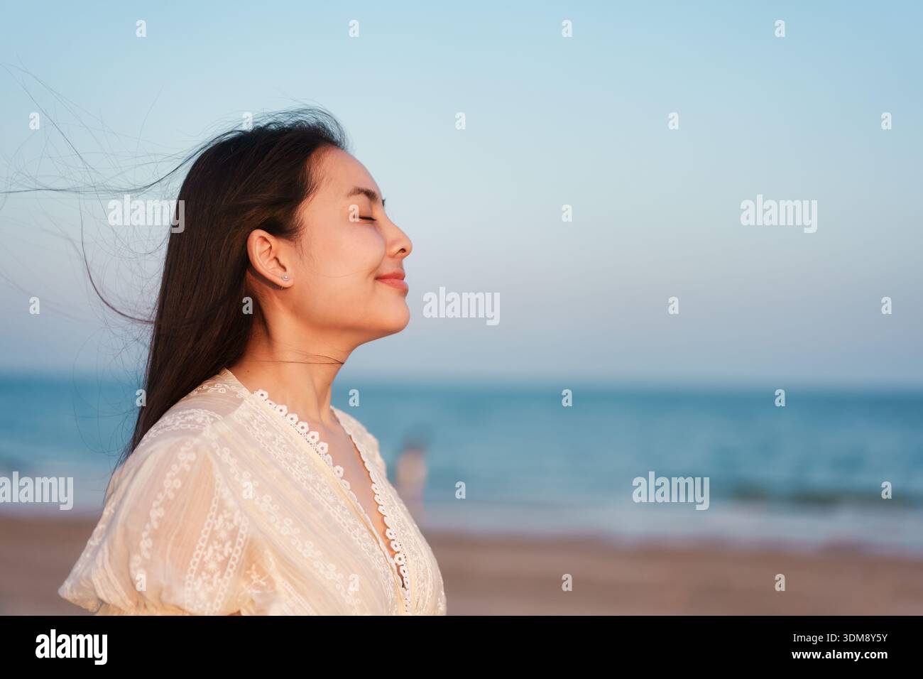 Ruhige asiatische Frau, die am Strand steht und tief in sanftem Abendlicht einatmet und Entspannung, Achtsamkeit, emotionales Gleichgewicht und geistige Heilung ausdrückt Stockfoto