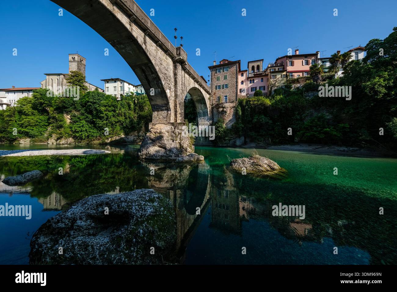 Die Steinbrücke Devil's Bridge überspannt den türkisfarbenen Natisone River mit der Kirche San Francesco und alten Häusern in der Ferne. Stockfoto